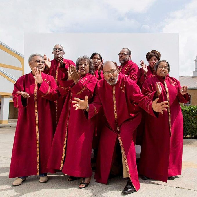 Group in red robes singing enthusiastically outdoors, with a church-like building in the background under a partly cloudy sky.