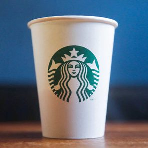 A white paper cup sits on a wooden table with a blue background, featuring a green mermaid logo in the center.