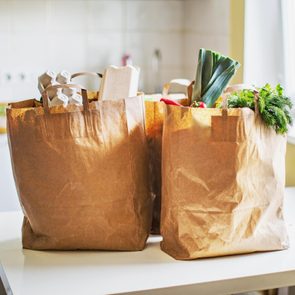 Brown paper bags sit filled with groceries, including vegetables and cartons, on a white tabletop in a bright kitchen.