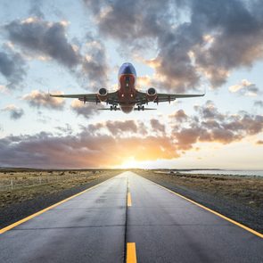 Airplane ascending over straight runway during sunrise, surrounded by open fields and a cloudy sky.