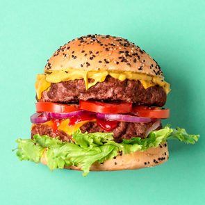 Burger displaying layers of beef, cheese, lettuce, tomato, and onions, sits against a solid teal background.