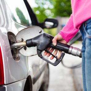 Hand holds gas pump nozzle refueling car at a station, wearing jeans and pink sweater, with trees in the background.