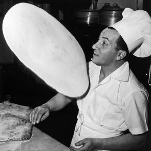 Chef tosses pizza dough into the air, focusing in a bustling kitchen environment.
