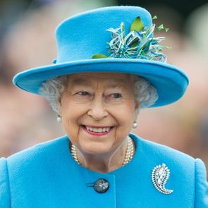 A person smiles while wearing a vibrant blue hat and coat, adorned with a brooch and pearls, amidst a blurred outdoor background.