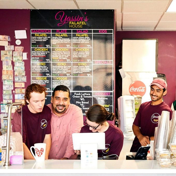 Counter staff chatting, smiling, and taking an order in a falafel restaurant with a menu board and Coca-Cola dispenser.