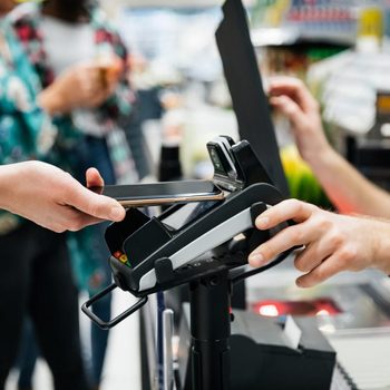 Hand holds smartphone near payment terminal, completing a transaction. Background shows blurred figures in a store setting.