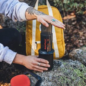Person presses Aeropress coffee maker on rock beside a yellow backpack in an outdoor setting.
