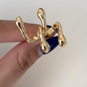 Gold, abstract ring entwines two fingers, held by a hand with blue nail polish against a plain background.