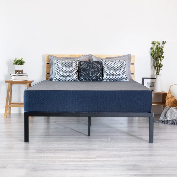 Bed with patterned pillows rests on a modern frame, surrounded by a side table with books and a plant in a bright, minimalist room.