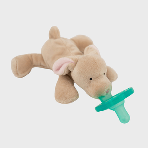 A beige plush elephant holds a green pacifier in its mouth, lying on a flat, white background.
