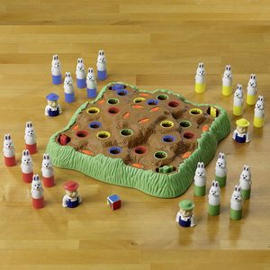 Plastic rabbits surround a carrot patch game board, with colorful holes, on a wooden table, accompanied by farmers and a small cube.