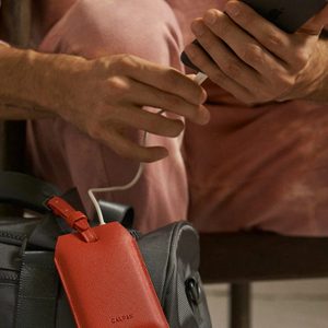 Hand plugs smartphone charger into gray bag with red tag, sitting on a bench.