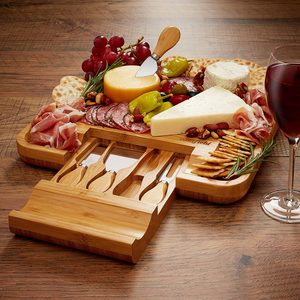 Cheese board displays various cheeses, meats, and grapes on a wooden surface with a drawer of knives and a glass of red wine nearby.