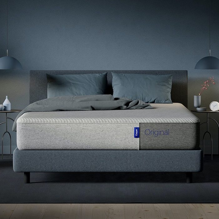 A neatly made bed with gray sheets and pillows rests between two side tables in a dimly lit bedroom. Text: 
