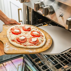 Pizza, topped with tomatoes and cheese, is being placed into a stainless steel oven on a wooden paddle in a kitchen setting.