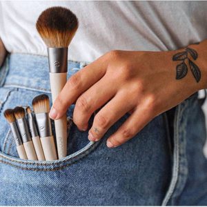 Makeup brushes tucked in jean pocket; hand adjusting, tattooed arm nearby.