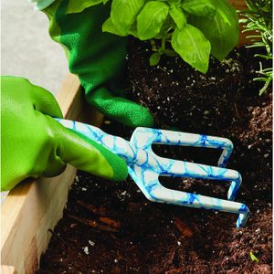 Hand uses blue-patterned garden fork to loosen soil around green plant in a wooden planter, wearing bright green gloves.