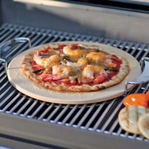 Pizza topped with shrimp bakes on a round stone inside a grill, surrounded by grilled vegetables on the side.