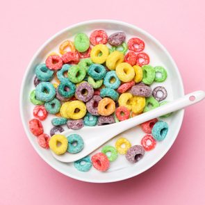 A bowl contains colorful ring-shaped cereal with a spoon, set against a pink background.