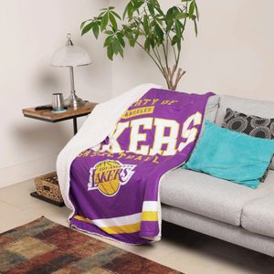 Blanket draped on a couch, displaying Los Angeles Lakers logo, surrounded by cushions, side table with lamp, potted plant, and a rug.