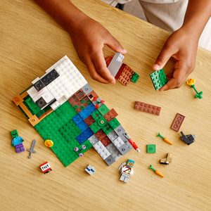 Hands build with colorful LEGO pieces on a wooden table; various blocks and small figures scattered.