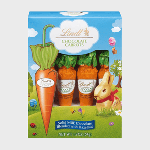 Packaging displays chocolate carrots with hazelnut, surrounded by a scenic landscape with flowers, clouds, a hot air balloon, and a gold bunny.