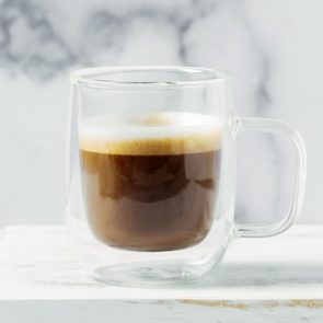 A clear glass mug holds frothy coffee, resting on a white surface with a light, blurred background.
