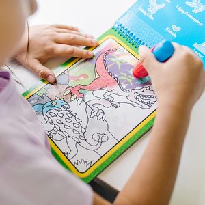 Child colors dinosaur illustration using water pen on spiral-bound coloring book at a table.