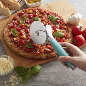Pizza cutter poised over vegetable-topped pizza on wooden board, surrounded by tomatoes, garlic, basil, and shredded cheese on a countertop.