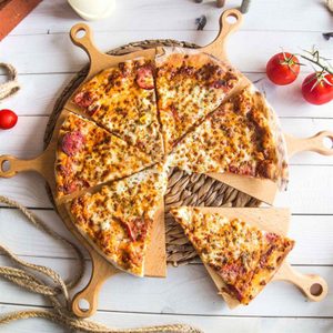 Pizza sliced on wooden boards, resting on a woven mat, surrounded by rope and fresh tomatoes, on a white wooden table.