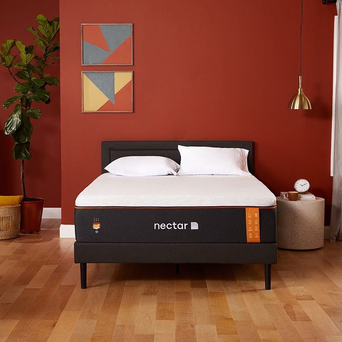 Bed stands in cozy bedroom; pillows resting; adorned with modern art, potted plant, and side table. Text reads 