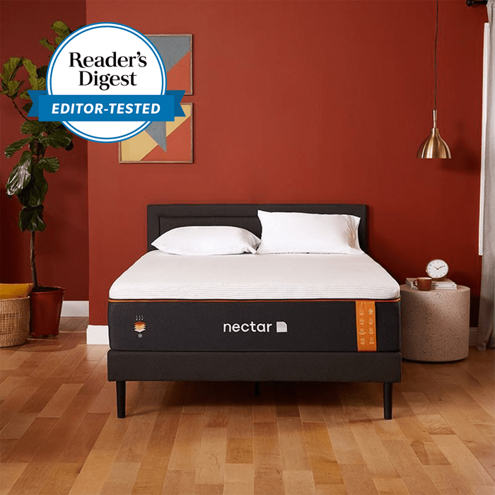 Bed stands on wooden floor, adorned with white pillows, against a red wall; potted plant, pendant lamp, and geometric artwork nearby. 