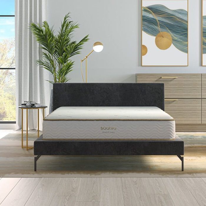 Bed with sleek mattress sits beside a gold lamp; modern room features plants, artwork, and natural light from large window. 