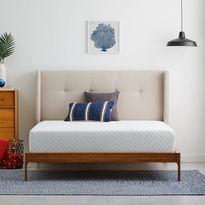 Bed with cushions sits in a bedroom; surrounded by a dresser, potted plant, wall art, hanging lamp, and carpet on tiled floor.