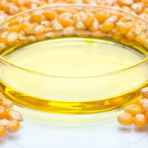 Yellow oil sits in a clear glass dish surrounded by scattered corn kernels on a white surface.