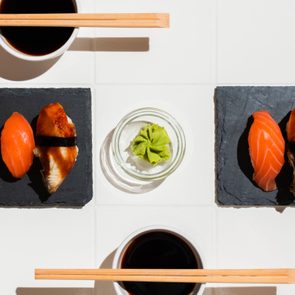 Sushi arranged on black slates, surrounded by soy sauce cups and chopsticks on a white tabletop. A small dish holds green wasabi.