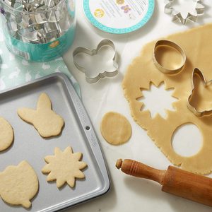 Cookie cutters shape dough into butterfly and flower forms on a counter, while baked cookies rest on a tray beside a rolling pin.