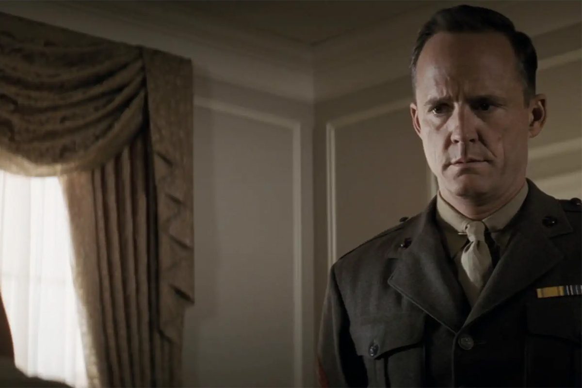 A man in a military uniform stands solemnly inside a room with elegant draped curtains.