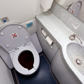 Toilet with open lid, no smoking sign, beside a sink in an airplane lavatory. Tissue holder and faucet detail visible.