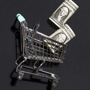 Miniature shopping cart holds rolled dollar bills against a dark background.