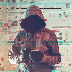 A hooded figure holds a smartphone, obscured by digital glitches, against a newspaper-covered wall.