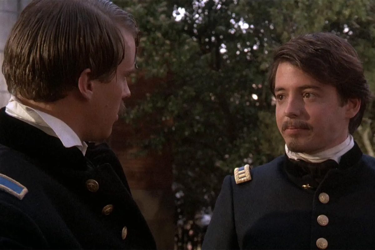 Two men in military uniforms converse outdoors, surrounded by trees.