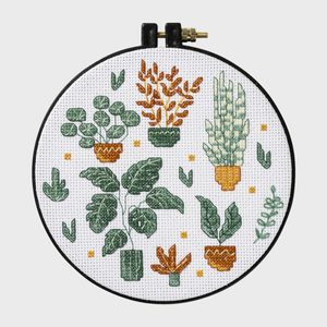 Embroidery hoop displays cross-stitched green and orange plants, surrounded by small leaves and dots.