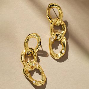 Gold earrings dangle elegantly with interlinked loops accented by small diamonds, resting on a textured beige surface.