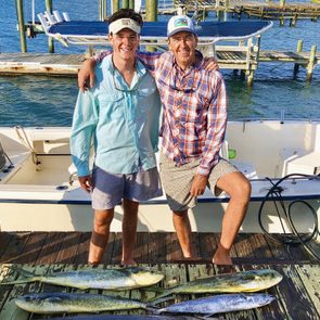 Two people smile on a dock beside a boat, displaying freshly caught fish, with a marina in the background under a sunny sky.