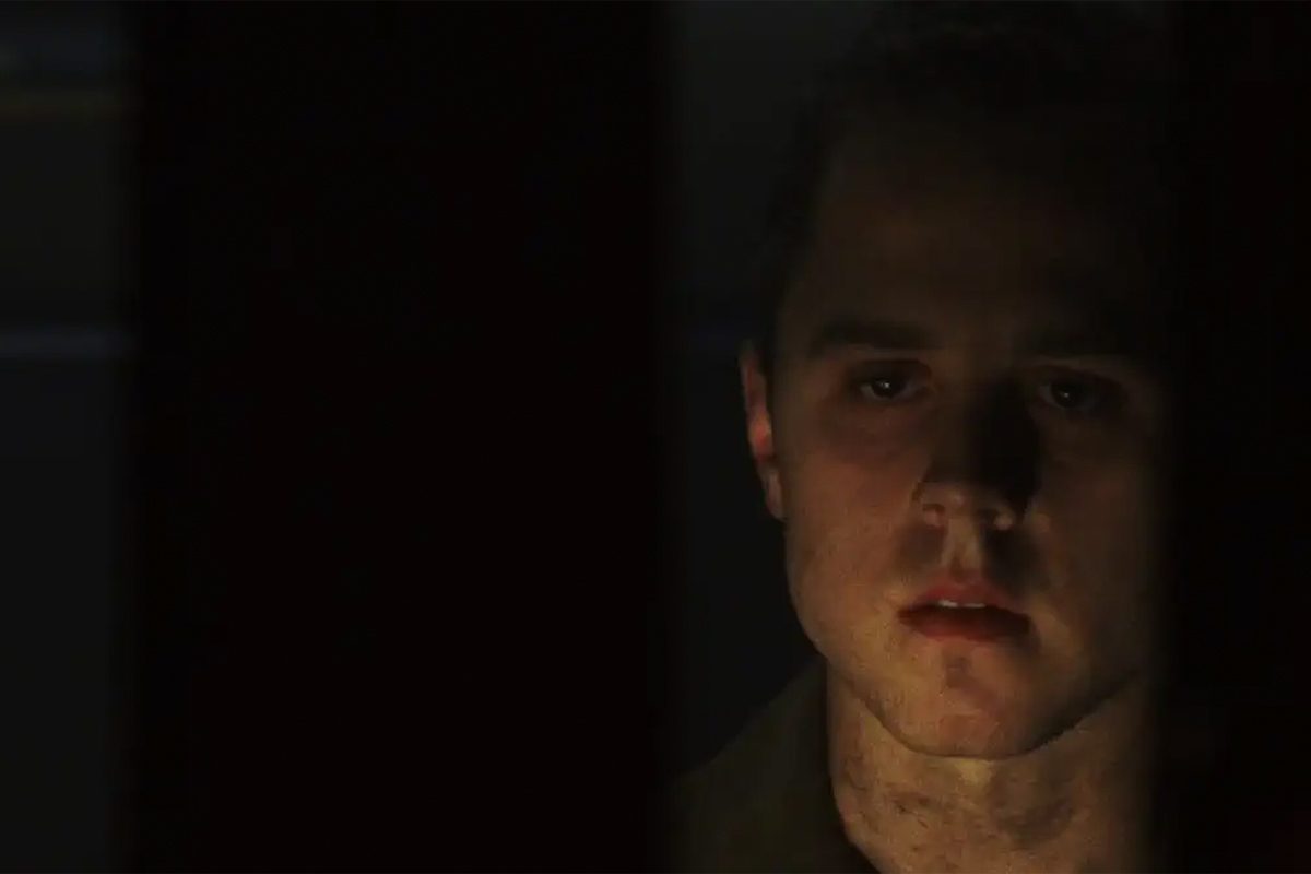 A person's face partially illuminated, emerging from shadows, with a contemplative expression against a dark background.