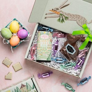Chocolate rabbit and carrot cake bar sit in a gift box with wrapped candies, next to colorful eggs in a pastel setting.
