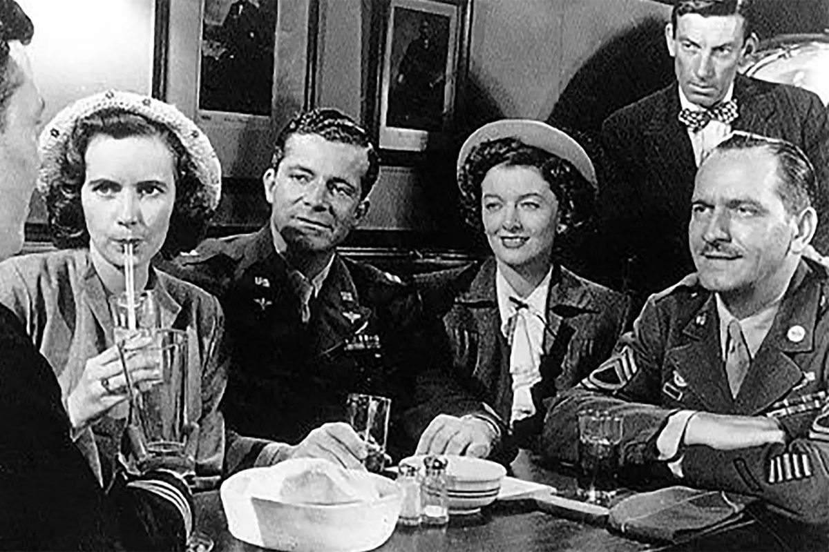 Five people sit at a diner table, conversing and sipping drinks, wearing 1940s attire, with military uniforms and hats, surrounded by framed photos.