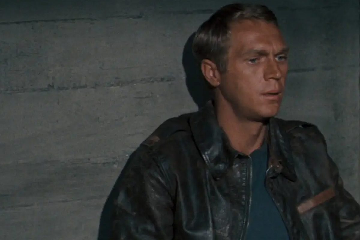 Person sits against a concrete wall, wearing a leather jacket, with a neutral expression. The background is plain and dimly lit.
