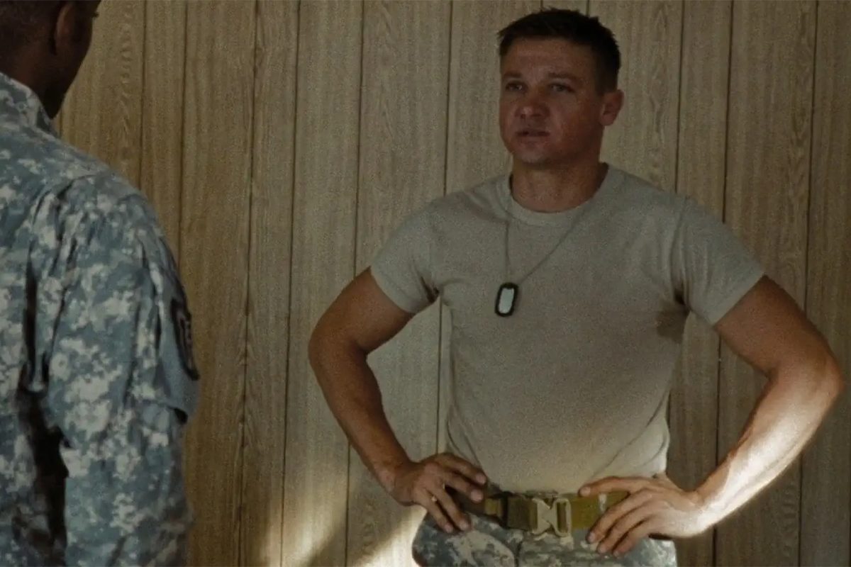 Man in military uniform stands hands on hips, facing another person, against a wooden wall backdrop.